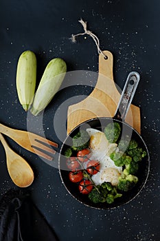 Healthy breakfast: broccoli, cherry tomatoes and fried eggs in a pan, which stands on a wooden stand on a dark background.