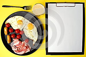 A healthy breakfast - coffee, scrambled eggs, bacon, cheese and cherry tomatoes. Food in a black plate on a yellow background.