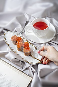 Healthy breakfast concept with a red caviar pancakes in female hand and cup of fruit tea in bed. Open book lie nearby.