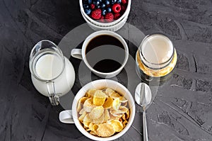 Healthy breakfast with cornflakes in a white cup, berries, milk and coffee on a dark gray background.