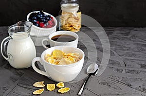 Healthy breakfast with cornflakes in a white cup, berries, milk and coffee on a dark gray background.