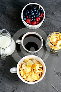 Healthy breakfast with cornflakes in a white cup, berries, milk and coffee on a dark gray background.