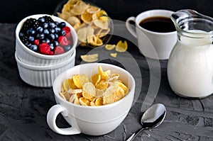 Healthy breakfast with cornflakes in a white cup, berries, milk and coffee on a dark gray background