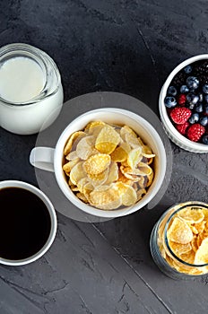 Healthy breakfast with cornflakes in a white cup, berries, milk and coffee on a dark gray background