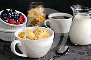 Healthy breakfast with cornflakes in a white cup, berries, milk and coffee on a dark gray background.