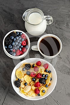 Healthy breakfast with cornflakes in a white plate, berries, milk and a cup of black coffee on a dark gray background