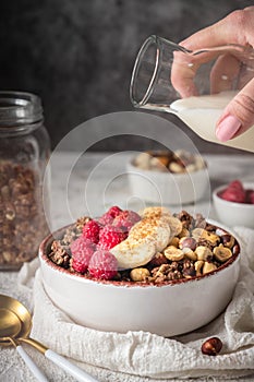 Healthy breakfast granola in a plate with nuts, banana and raspberries, milk is poured from a bottle