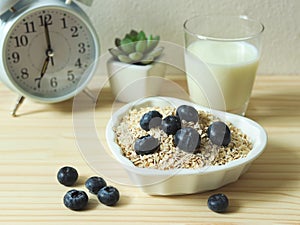 healthy breakfast with oat or granola in heart shape bowl, fresh blueberries, milk and white alarm clock 7 am on wooden table.