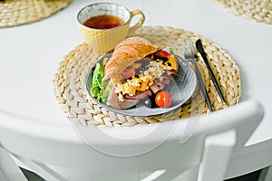 Healthy breakfast. Plate with croissant sandwich with ham, scramble egg, tomato with tea on table