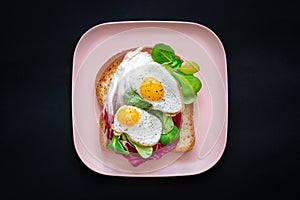 Healthy breakfast sandwiches concept. Bread toasts with fried eggs and green salad on pink plate on black