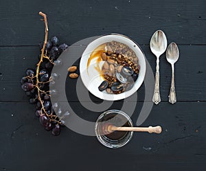 Healthy breakfast set. Bowls of oat granola with yogurt, fresh grapes, almond and honey over black wooden backdrop