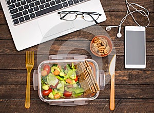 Healthy business lunch at workplace. Salad, salmon, avocado and nuts lunch box on working desk with laptop, smartphone, glasses