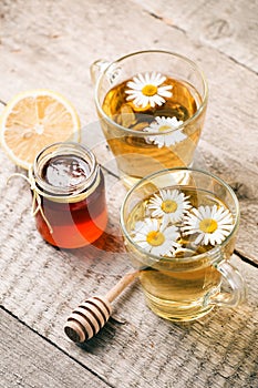 Healthy chamomile tea poured into glass cup. Teapot, small honey jar, heather bunch and glass jar of daisy medicinal herbs