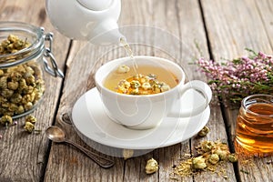 Healthy chamomile tea poured into white cup. Teapot, honey jar, heather bunch, jar of daisy herbs.