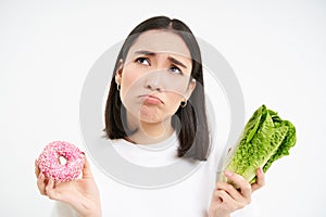 Healthy clean detox eating concept. Vegetarian, vegan, raw concept. Sad asian girl on diet holds doughnut and vegetable