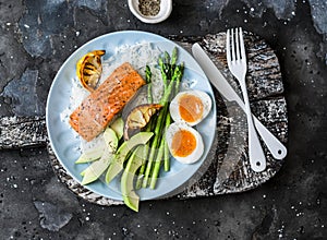 Healthy delicious balanced lunch - baked salmon, rice, asparagus, avocado and boiled egg on a dark background, top view