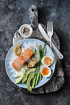 Healthy delicious balanced lunch - baked salmon, rice, asparagus, avocado and boiled egg on a dark background, top view