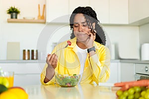 Healthy diet. Sad african american woman eating fresh vegetable salad, sitting at dinner table in kicthen, free space