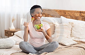 Healthy diet for weight loss concept. Happy black woman eating yummy vegetable salad on bed at home, blank space