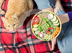 Healthy dinner or lunch. Woman in jeans sitting on red blanket with crossed legs and eating vegan in a bowl with vegetables,