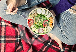 Healthy dinner or lunch. Woman in jeans sitting on red blanket with crossed legs and eating vegan in a bowl with vegetables,