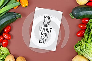 Healthy eating concept with `You are what you eat` text card on brown background with salad, zucchini, tomatoes, potatoes and bell