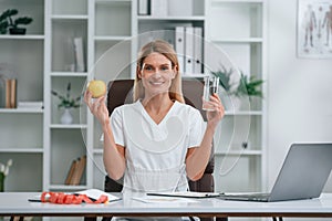 Healthy eating, holding an apple and glass of water. Conception of diet. Young female doctor in white coat is indoors