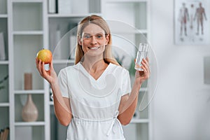 Healthy eating, holding an apple and glass of water. Conception of diet. Young female doctor in white coat is indoors