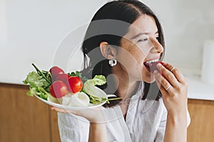 Healthy eating. Young happy woman eating cherry tomato and  holding plate with lettuce, tomatoes