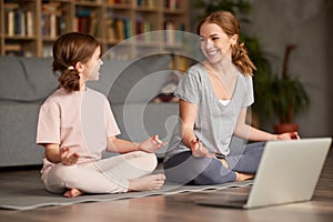 Healthy family mom and daughter meditating together at home while having online yoga class on laptop