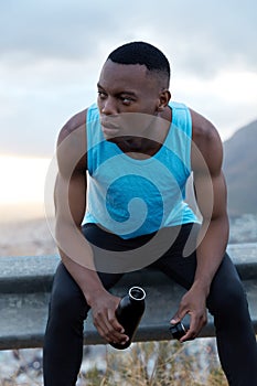 Healthy fatigue dark skinned man takes break after outdoor workout, holds bottle of energy drink, has pensive look