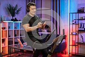 Healthy fit smiling asian man training at home on exercise static bike during workout holding phone