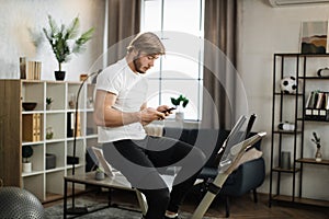 Healthy fit smiling asian man training at home on exercise static bike during workout holding phone