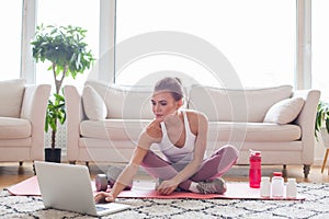 Healthy fit young woman in activewear sitting on mat with laptop, water and protein bottles at home