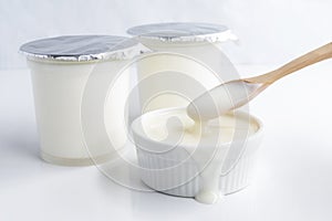 Healthy flavored yogurt in plastic cup and ceramic bowl with wooden spoon on the white table background