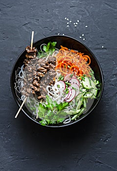 Healthy food bowl - beef skewers, rice noodles and vegetable salad on dark background