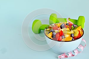 Healthy food. Bowl of vegetarian fresh fruit salad made of different fruits, dumbbells, measuring tape: banana, kiwi, apple,