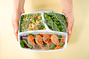 Healthy food in lunch box in female hands top view. Shrimps, rice, salad, arugula and vegetables are in the delivery container.