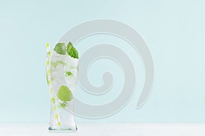 Healthy fresh tonic with green mint, ice cubes, soda, striped straw in misted glass on white wood table, pastel green color.