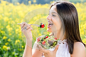 Healthy girl eating salad outdoors with flower field in background.