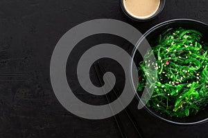 Healthy green seaweed salad with sesame seeds in a black bowl, peanut sauce and chopsticks on a slate black background.