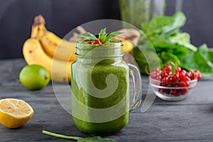 Healthy green spinach smoothie in a jar mug decorated with mint and red currant berries with ingredients on the black wooden table