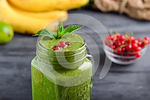 Healthy green spinach smoothie in a jar mug decorated with mint and red currant berries with ingredients on the black wooden table