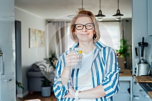 Healthy habit to drink water. Smiling middle age woman with glass of pure water with lemon standing on her kitchen