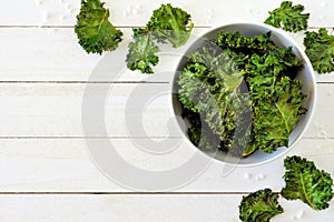 Healthy kale chips in a bowl, above scene on white wood with copy space.