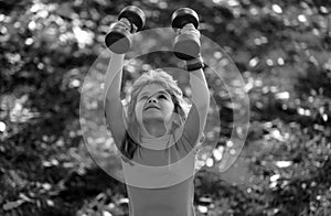 Healthy kid boy with dumbbell exercise outdoor. Little child practice dumbbells exercises in park. Funny excited child