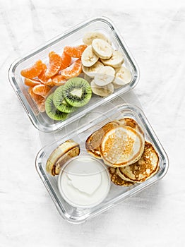 Healthy kids school lunch box - pancakes with sour cream and banana, kiwi, tangerine fruit. Delicious snack on light background