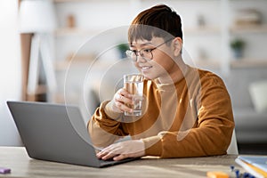 Healthy korean teenager drinking water while using laptop at home