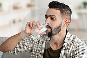Healthy lifestyle concept. Millennial Arab guy drinking water from glass at home