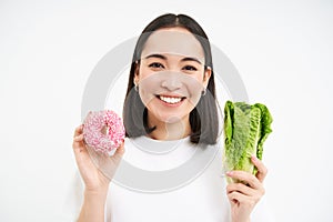 Healthy lifestyle concept. Smiling asian woman shows doughnut and cabbage, choice of eating unhealthy junk food or